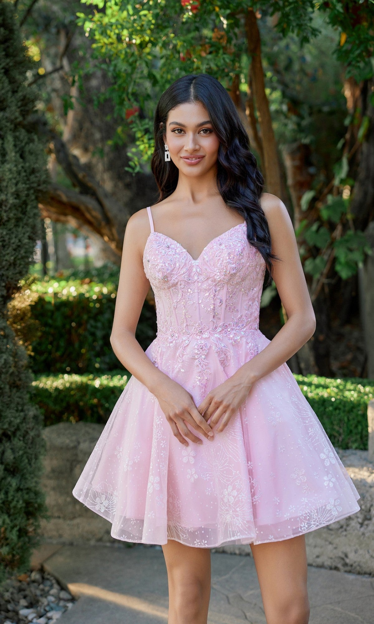 A woman stands outdoors in a garden, wearing the Juliet JT985L short homecoming dress in blush, featuring a sleeveless design with floral details and a delicate corset bodice.
